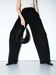 Model wearing black wide leg trousers and a white top holding a black leather handbag against a plain background