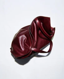 Burgundy leather shoulder bag resting on a white seamless background with soft directional shadow casting beneath it.