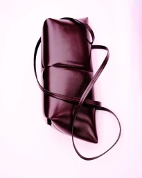 A structured dark burgundy leather handbag with a thin shoulder strap resting against a pale pink background.