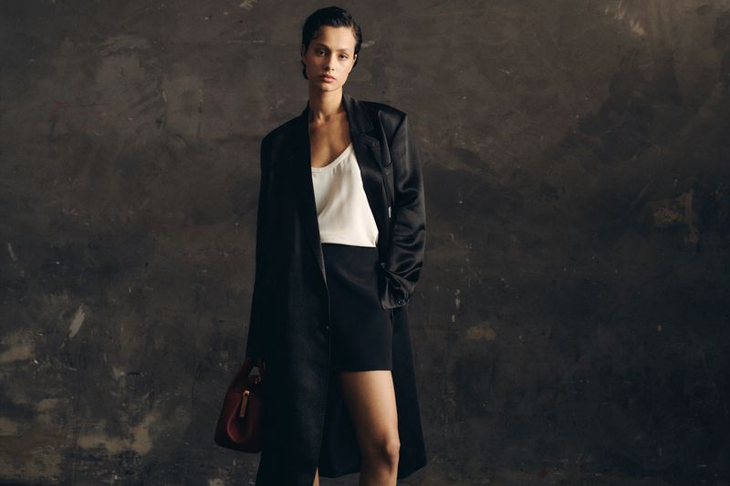 Fashionable woman in black coat and shorts stands against a textured dark gray wall holding a purse, creating a sleek, modern aesthetic.