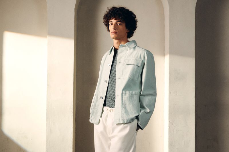 Adult male with curly hair posing in front of a wall in light blue jacket and white pants, standing in a light-filled arched doorway.