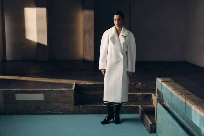 Elegant woman posing in a long white coat in a minimalist setting, bathed in natural light with contrasting dark shadows and blue accents.