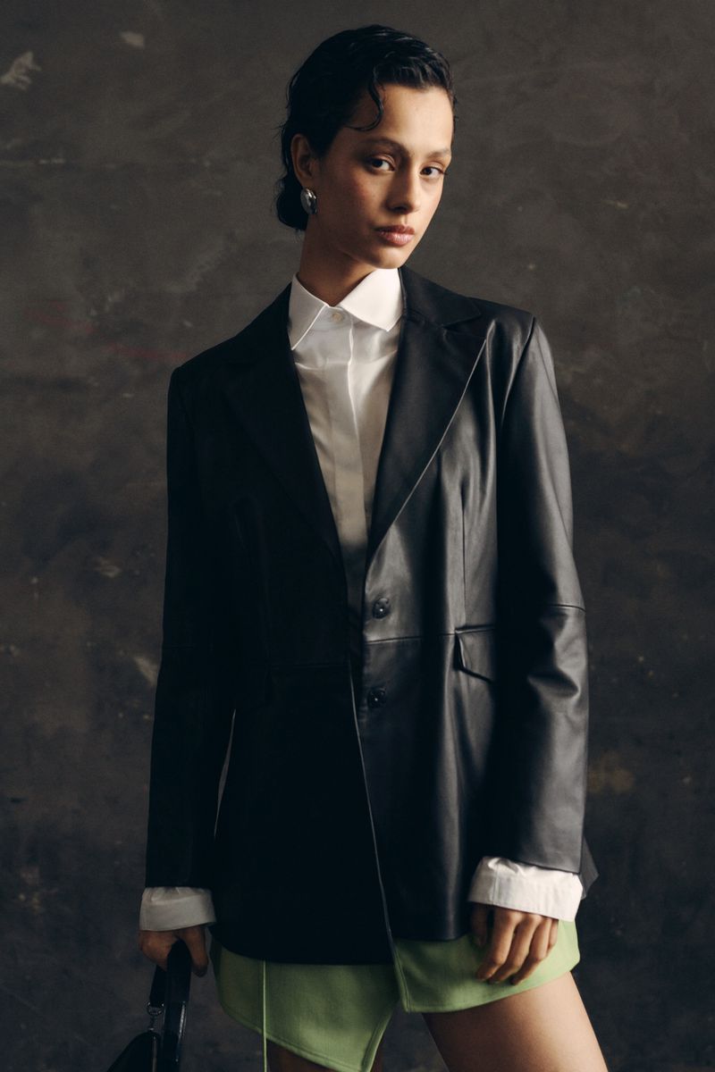 Woman in a black leather blazer and light green skirt posing in front of a textured dark grey backdrop in a fashion photography shot.