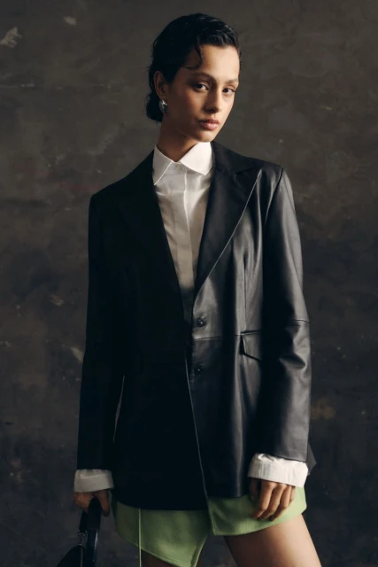 Woman in a black leather blazer and light green skirt posing in front of a textured dark grey backdrop in a fashion photography shot.