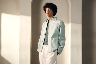 Male model with curly dark hair wearing a light blue jacket and white trousers against an arched architectural backdrop.