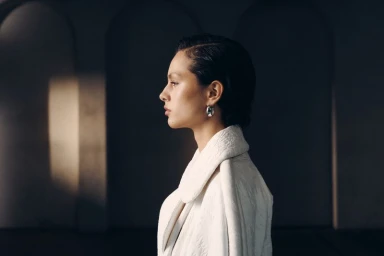 Profile portrait of a woman wearing a white textured coat and silver hoop earring against an arched architectural background.