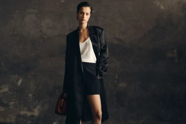 Model wearing a black coat and white top holding a dark red handbag standing before a textured dark wall.