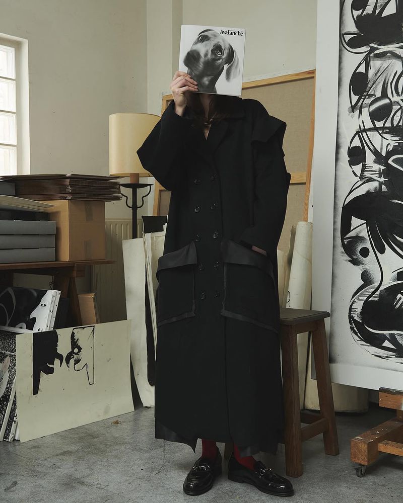 Woman in black coat holds record with dog face in art studio, surrounded by paintings and supplies, creating a fashion-forward editorial scene.