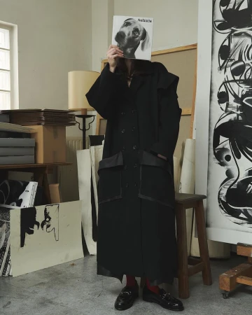 Woman in black coat holds record with dog face in art studio, surrounded by paintings and supplies, creating a fashion-forward editorial scene.