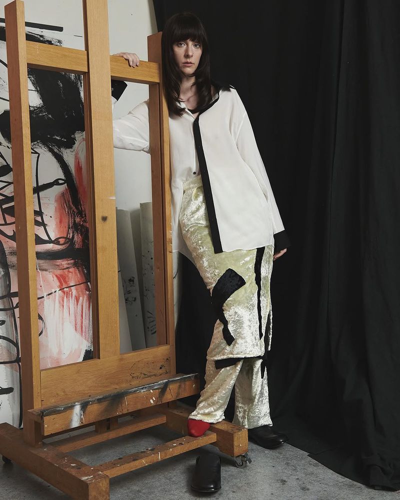 Fashionable adult female poses in a white blouse and textured pants next to a painted easel in a studio with black backdrop.