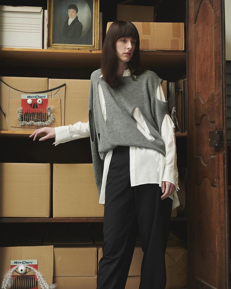 A woman in a gray sweater with cutouts poses indoors against a wooden shelving unit filled with boxes and a framed portrait.