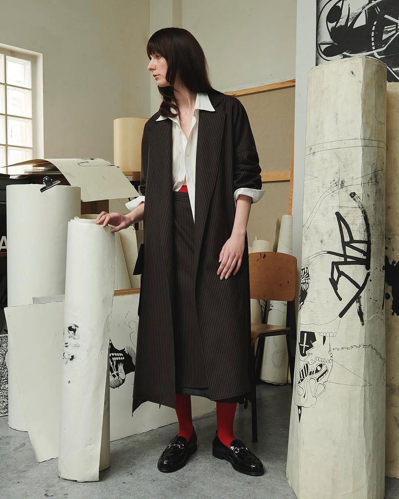 A stylish woman in a pinstripe coat, red tights, and loafers stands in a light-filled artist studio, surrounded by large paper rolls with drawings.