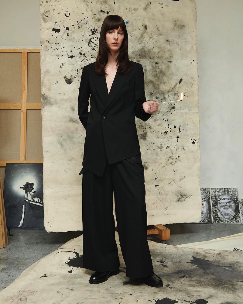 A long-haired adult stands in an art studio, dressed in black suit separates, holding a lit stick, with paintings in the background.