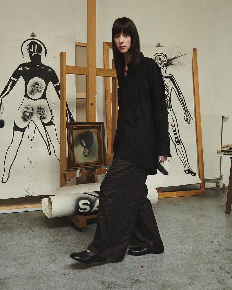 A woman in an oversized black coat and wide-leg pants poses in an artist's studio surrounded by drawings and art supplies.