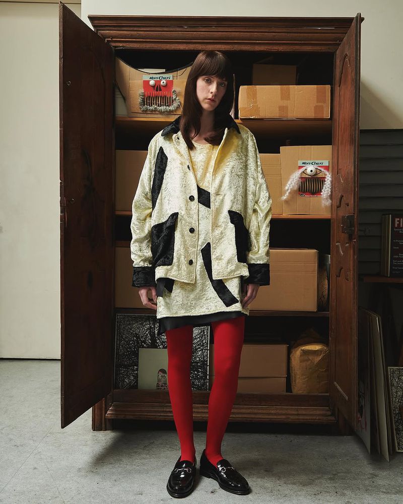 Indoor portrait of a woman in patterned jacket and dress with red tights, standing in front of an open wooden wardrobe cabinet.