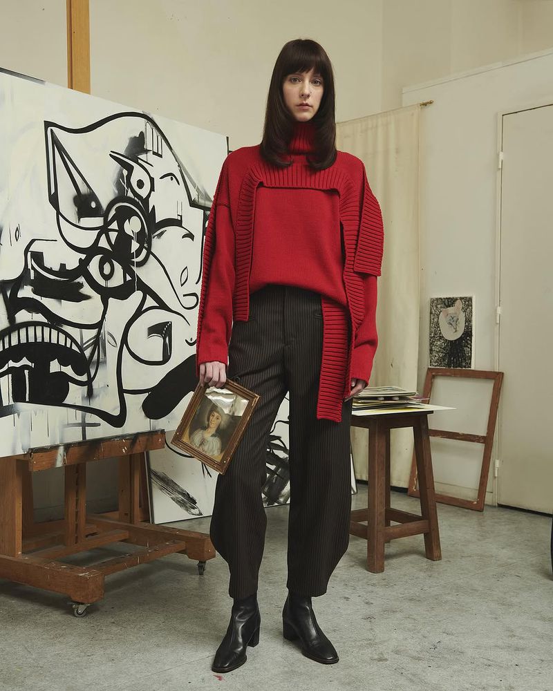 A slender woman in a red sweater, striped pants, and black boots holds a framed picture in an artist studio with abstract art.