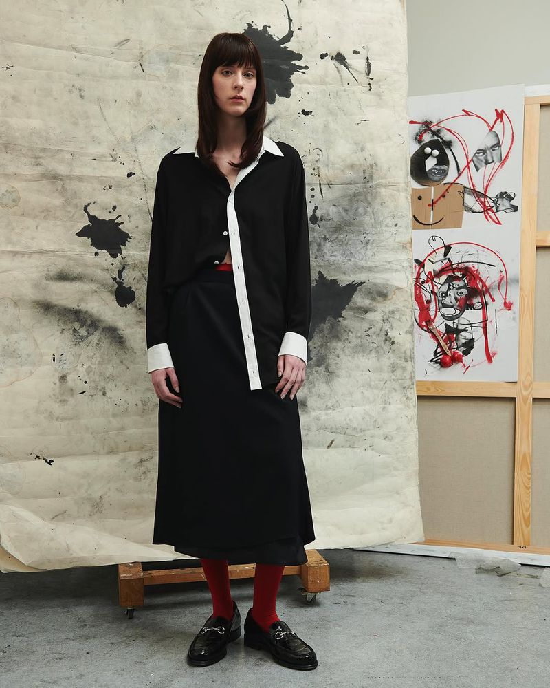 Adult female in black-and-white collared shirt, black skirt, and red tights stands in front of a painted backdrop in a studio.