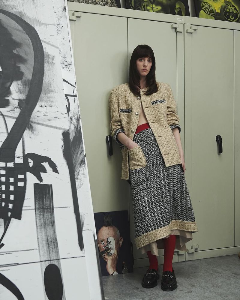 A woman in a beige jacket and patterned skirt poses next to abstract artwork indoors. She has her hand in her pocket and is looking forward.