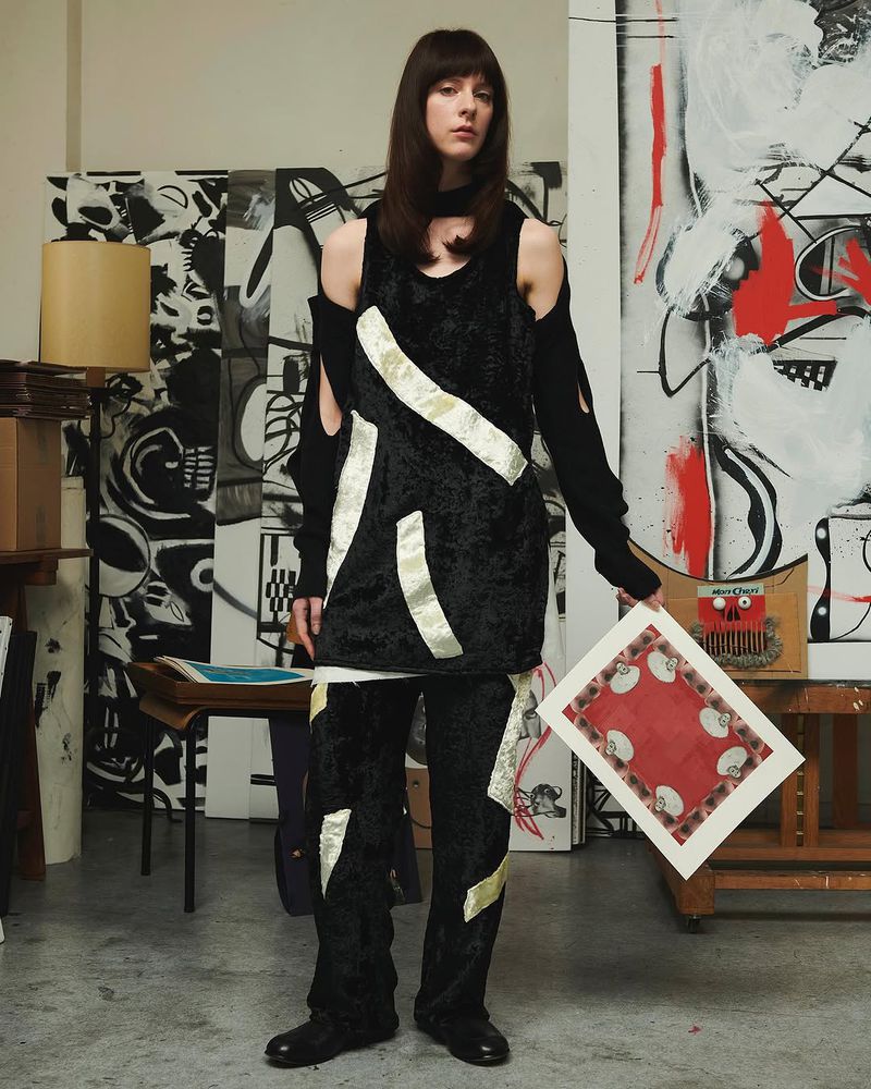 A woman in a black outfit poses in an artist's studio, holding a framed piece of art in front of a painted background with mixed media.