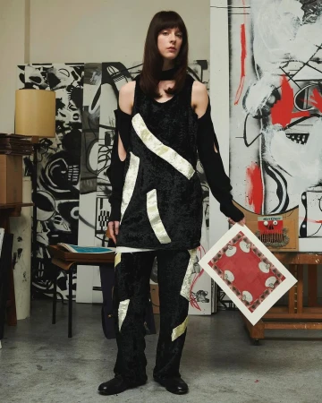 A woman in a black outfit poses in an artist's studio, holding a framed piece of art in front of a painted background with mixed media.