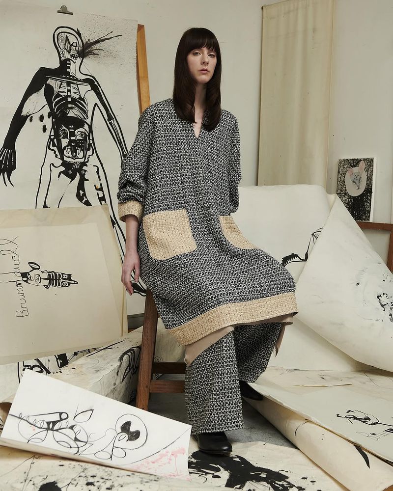 Female fashion model sits on a wooden stool amid paper art and wears patterned grey tunic with tan pockets and matching trousers.