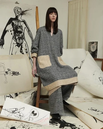 Female fashion model sits on a wooden stool amid paper art and wears patterned grey tunic with tan pockets and matching trousers.