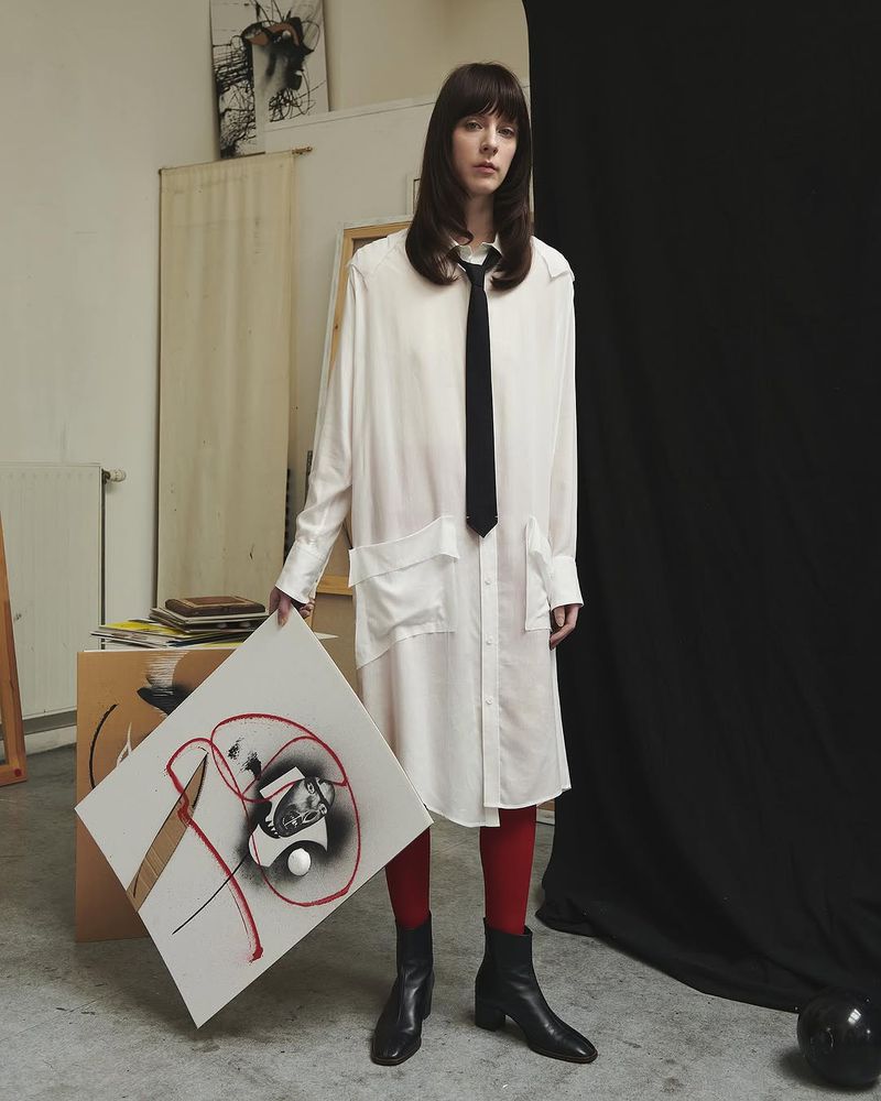 Adult female in a white dress shirt with a black tie and red tights, holding an abstract painting inside an art studio.