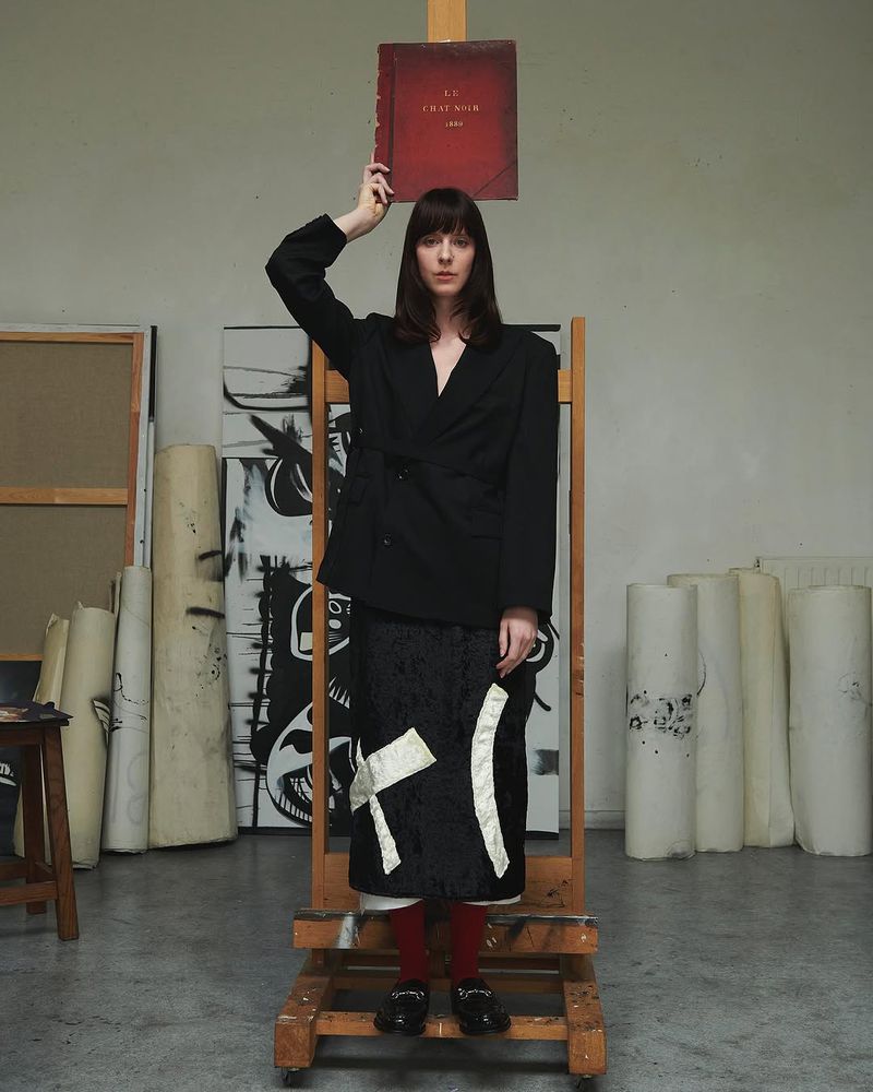 A woman in a black blazer and skirt stands in an artist studio, holding a red book above her head in a creative, artistic setting.