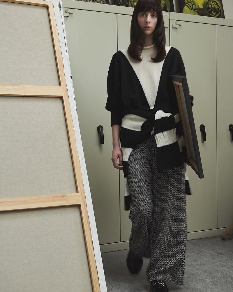 Indoor shot of a woman in patterned pants and a black and white top carrying a framed painting near a large canvas and lockers.