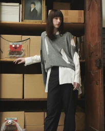 Model standing in a room with cardboard boxes wearing a grey sleeveless knit top over a white shirt and black trousers.
