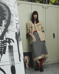 Model leaning against a pale storage cabinet wearing a textured beige jacket and patterned skirt in an interior setting