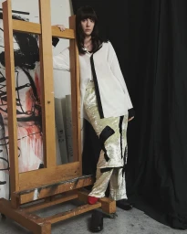 Model leaning against a large wooden artist easel wearing a white shirt and patterned trousers in an artist studio.
