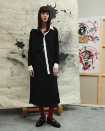 Model standing in a black skirt and matching top with white collar and cuffs against an artistic studio backdrop