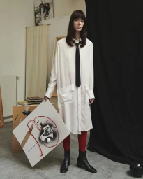Model standing in an art studio wearing a white shirt dress, black tie, red tights, and black boots, holding a framed painting.
