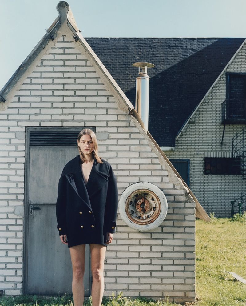 Fashion shot of a fair-skinned adult woman with long brown hair wearing a black double-breasted coat standing in front of a white building.