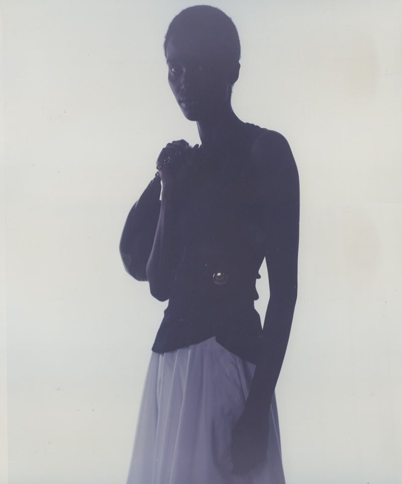 A person stands with a purse over their shoulder wearing a black top and white skirt against a white background in a studio shot.