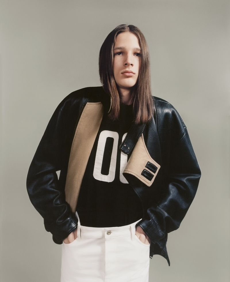Studio shot of a young adult male model wearing a black leather jacket over a sweatshirt, standing in front of a neutral background.