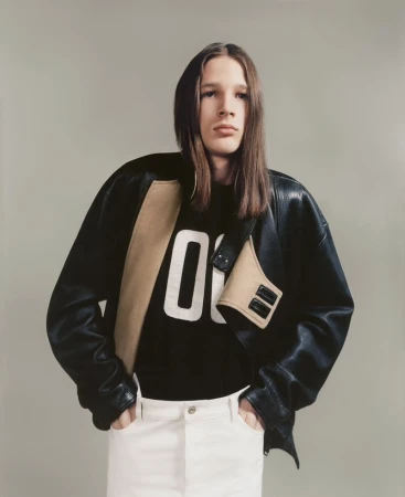 Studio shot of a young adult male model wearing a black leather jacket over a sweatshirt, standing in front of a neutral background.