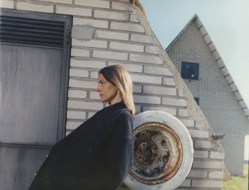 Woman in black coat poses against brick building and weathered white tire in rural setting under blue sky. Minimalist outdoor portrait.