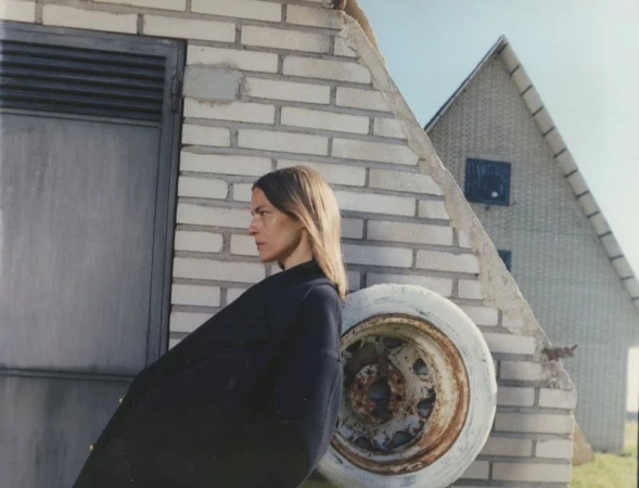 Woman in black coat poses against brick building and weathered white tire in rural setting under blue sky. Minimalist outdoor portrait.