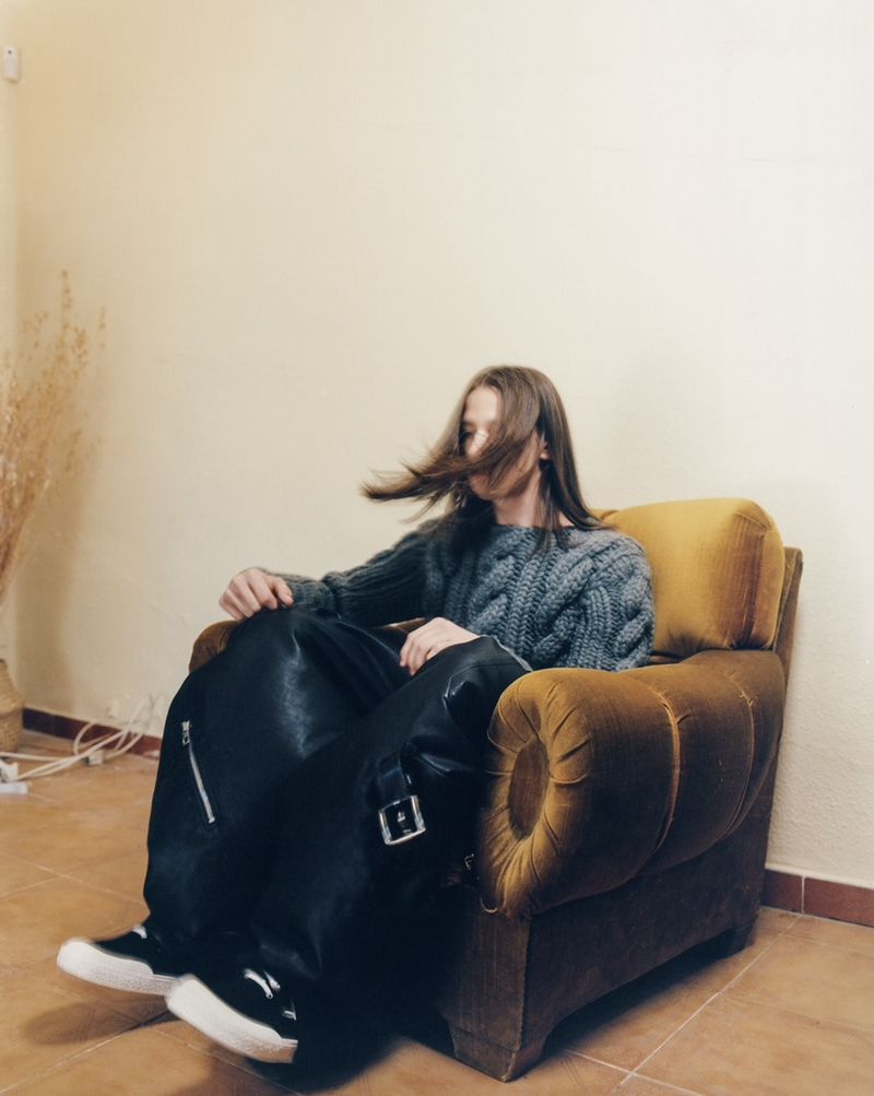 A woman with long brown hair, wearing a grey sweater, sits in an armchair with a black leather jacket draped across her lap in a home interior.