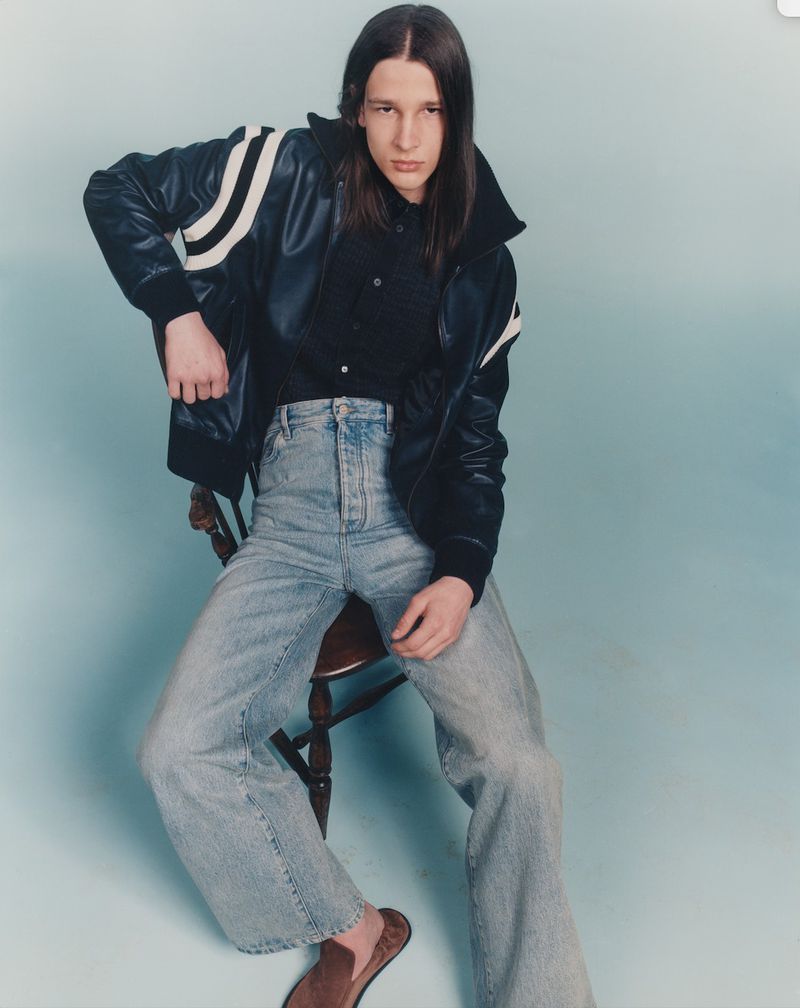 A young adult male with long brown hair sits on a wooden stool, wearing a bomber jacket, black shirt, light wash jeans, and brown loafers.