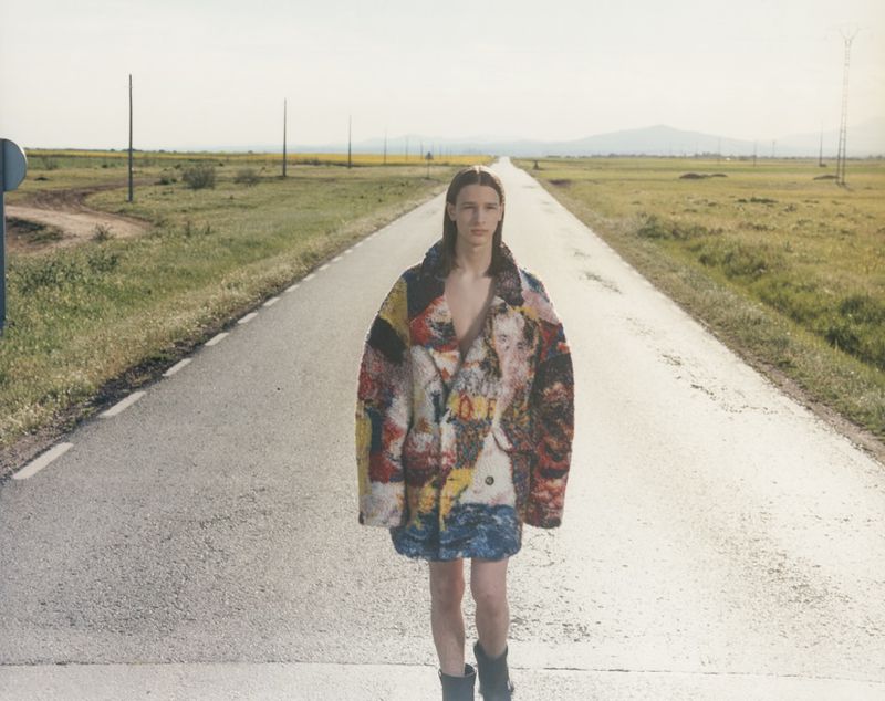 Fashion shot of a young adult wearing a colorful patterned coat walking down a rural road under a bright sky in a natural setting.
