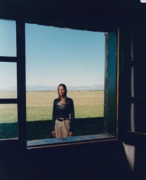 Model standing outdoors framed in a dark window view wearing a black jacket and tan trousers against a green field and blue sky.