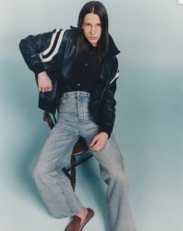 Model seated on a wooden chair wearing a dark leather jacket and light wash jeans against a blue gradient background.