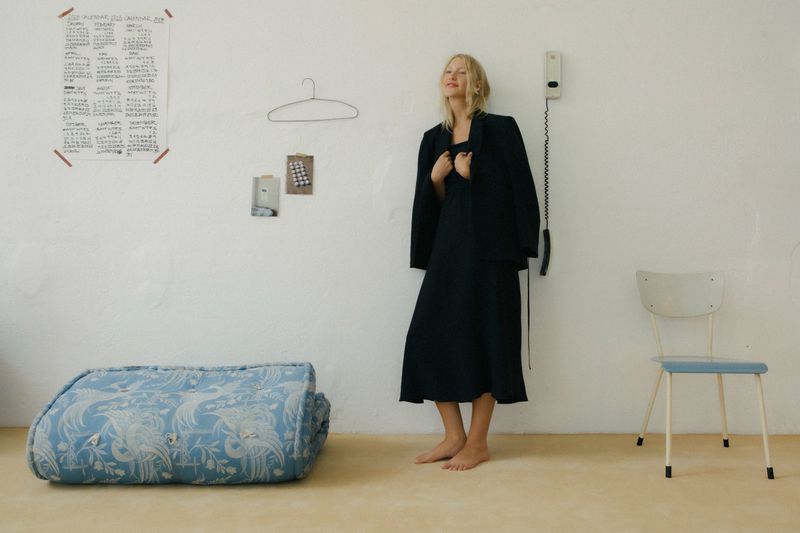 Blonde woman in a dress and blazer stands in an indoor setting next to a chair, with a patterned cushion on the floor.