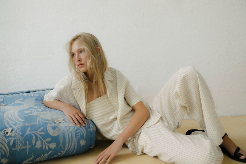 A fair-skinned woman reclines indoors wearing a beige striped linen suit and sandals, with a patterned cushion to her side.