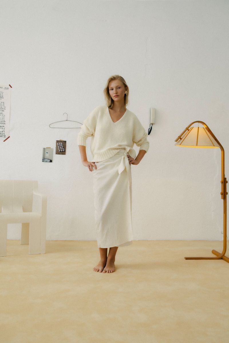 A blonde woman poses in a white sweater and wrap skirt, indoors, with cream-colored walls and floor, a lamp, and a white chair.