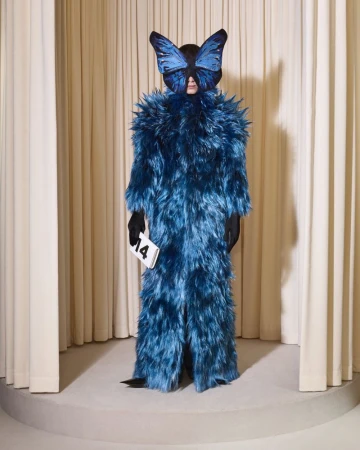 Person in a full-body blue furry costume and butterfly mask holding a card with '14' in front of a light beige curtain backdrop.