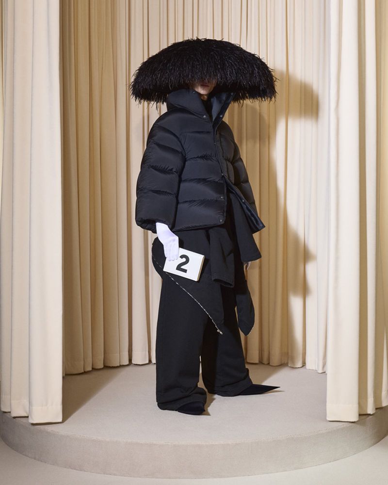 Person in black puffer jacket and wide-leg pants stands on a platform in front of draped beige curtains holding a card.